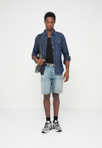 Chemise en denim, t-shirt noir, short bleu clair et baskets sportives. Sac à bandoulière à surface texturée, boucle de ceinture argentée, chaussettes noires.