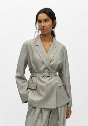 Femme portant un costume gris clair à fines rayures avec un blazer ceinturé et un pantalon large assorti, debout devant un fond blanc uni.