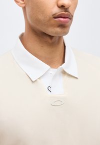 Κοντινή λήψη ενός ατόμου που φοράει πουκάμισο μπεζ χρώματος με γιακά πάνω από λευκό πουκάμισο με ορατή λεπτομέρεια μαύρου λογότυπου.