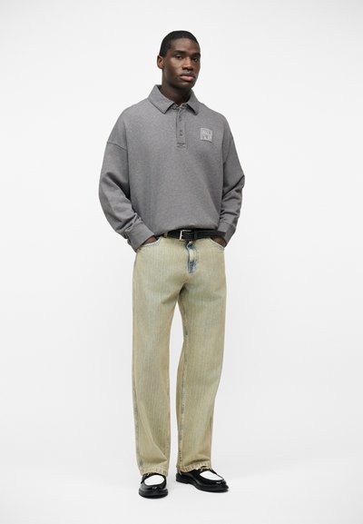 Άνδρας που φοράει γκρι μακρυμάνικο πόλο πουκάμισο, ανοιχτό μπεζ τζιν με μαύρη ζώνη και ασπρόμαυρα μοκασίνια, στέκεται με τα χέρια στις τσέπες.