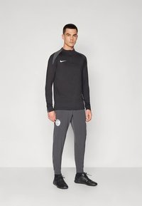 Zwart atletisch shirt met lange mouwen en een ritskraag, gecombineerd met grijze taps toelopende broek met een logo. De stof is glad en heeft een aansluitend ontwerp.