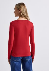 Rotes Langarmshirt mit Rundhalsausschnitt, aus weichem Material gefertigt. Verfügt über eine glatte Textur und eine lockere Passform, kombiniert mit blauen Jeans.