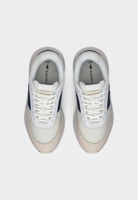 Πάνινα παπούτσια Lacoste με λευκό και ναυτικό μπλε χρωματικό συνδυασμό, λείο δέρμα, υφή από δίχτυ, στρογγυλή μύτη και επίπεδα κορδόνια. Χρυσό λογότυπο ως διακοσμητικό στοιχείο.