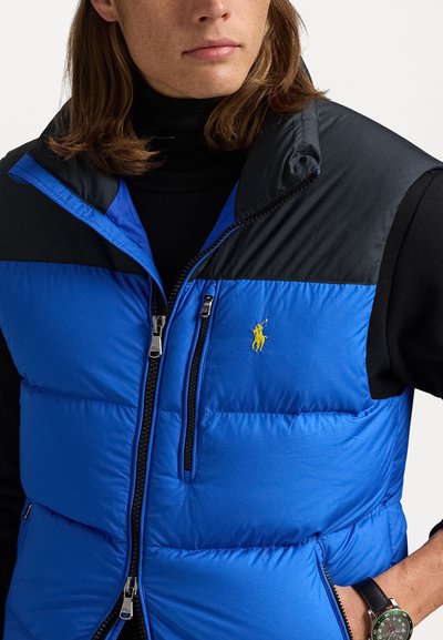 Zila un melna pūkaina veste ar kviltotu tekstūru, divām rāvējslēdzēju kabatām un dzeltenu logotipu uz krūškurvja. Valkāta virs melna augstvērtīga krekla.