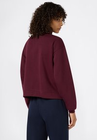 Felpa corta color bordeaux con maniche lunghe, colletto alto e polsini a costine. Presenta una vestibilità rilassata e una texture morbida.