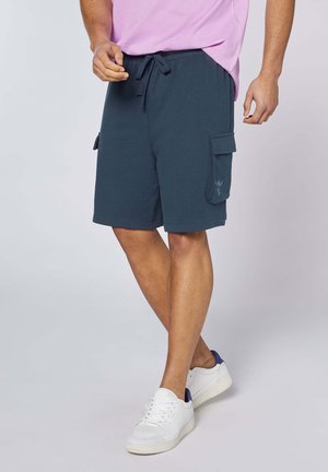 Mann trägt marineblaue Cargoshorts, pinkes Hemd und weiße Sneaker, steht mit einem Bein über das andere verschränkt vor einem einfarbigen Hintergrund.