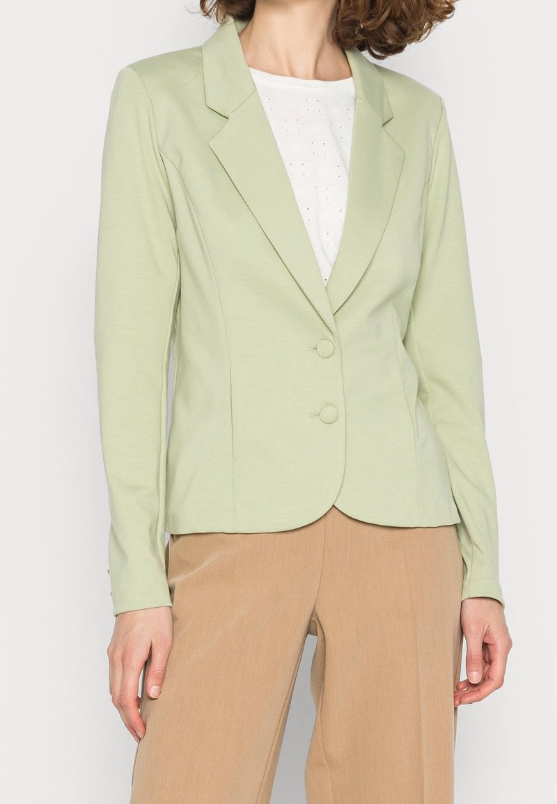 Femme portant un blazer vert clair à deux boutons sur un haut blanc et un pantalon beige, debout les bras détendus le long du corps.