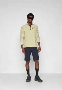 Pile verde chiaro con cerniera e maniche lunghe, abbinato a pantaloncini blu navy e scarpe da trekking nere. Modello con occhiali da sole neri.