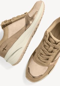 Zapatillas deportivas beige con una mezcla de materiales lisos y texturizados, que presentan un diseño de cordones, acentos metálicos y una suela de goma.