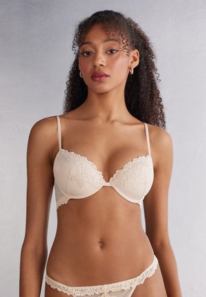 Beige padded bra with lace trim, featuring adjustable straps and a smooth texture. Subtle check pattern on fabric.