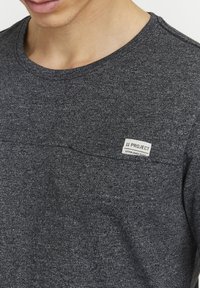 Graues Rundhals-T-Shirt aus strukturiertem Stoff, mit einem kleinen genähten Etikett auf der Brust mit "11 PROJECT" und "ESTABLISHED 2020."