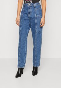 Jeans de ganga azul de cintura alta com pernas largas, apresentando dois bolsos laterais e costura em contraste. Combinados com botins de salto alto pretos.