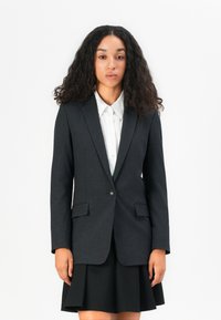 Upravená černá blazer s proužky, se dvěma předními kapsami, jedním zapínáním na knoflík a klopovými límci, nošená přes bílou košili s límečkem.