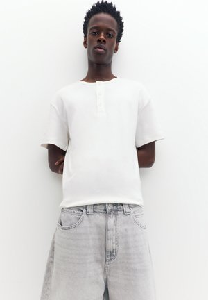 Jeune homme aux cheveux courts et bouclés portant une chemise blanche à manches courtes boutonnée et un jean gris clair sur un fond blanc uni.