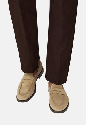 Jasnobrązowe mokasyny z zamszu noszone z ciemnobrązowymi spodniami, pokazujące zbliżenie dolnej części nóg i stóp.