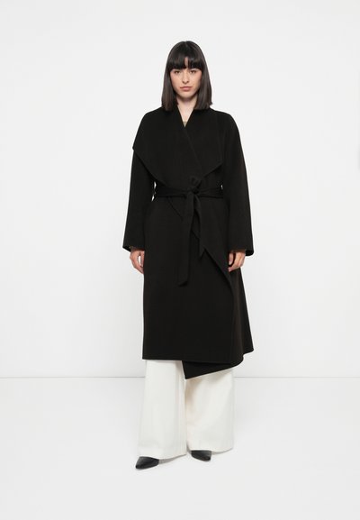 Cappotto avvolgente nero in misto lana con ampi rever, cintura da annodare e maniche lunghe, abbinato a pantaloni bianchi a gamba larga e tacchi neri.