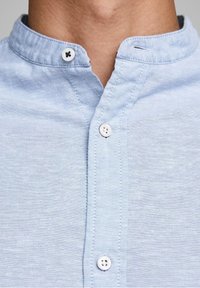 Ljusblå chambrayskjorta med mandarinkrage, två runda knappar vid halsen och subtil textur med sömnadsdetaljer.