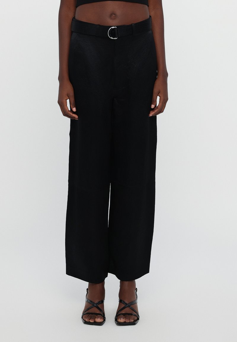 Zwarte broek met wijde pijpen en een gestructureerde afwerking, met een aansluitende tailleband en riem met een zilveren ronde gesp, gecombineerd met zwarte sandaaltjes met bandjes.
