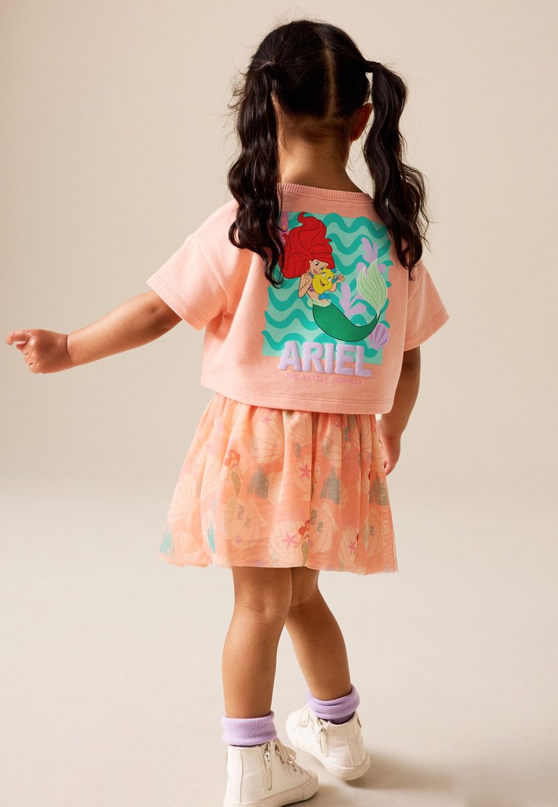 Niña joven con coletas que lleva una camiseta rosa con temática de Ariel, falda a juego con diseños de océano, calcetines morados y zapatillas blancas, de pie de lado.