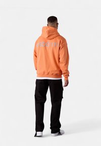 Haut à capuche orange avec "estéem" brodé dans le dos, associé à un pantalon cargo noir et des baskets blanches. Tissu lisse et coupe décontractée.