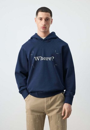 Joven con sudadera azul marino con la palabra "Where?" impresa y pantalones beige salpicados de pintura, de pie frente a un fondo liso.
