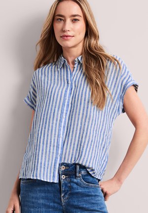 Frau mit langen hellbraunen Haaren, die ein kurzärmeliges blau-weiß vertikal gestreiftes Hemd mit Knöpfen und blaue Jeans trägt, stehend mit einer Hand auf der Hüfte.