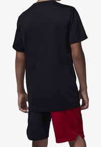Camiseta de algodón negra de manga corta, vista posterior. Combinada con pantalones cortos de malla rojos y negros que presentan un detalle de marca lateral.