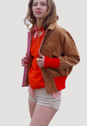 Veste en velours côtelé marron avec des poignets en ribbed orange, superposée à un pull orange et une chemise rayée, associée à un short crème.