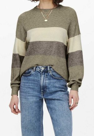 Kvinde iført en stribet sweater i taupe, creme og grå farver kombineret med højtsiddende blå jeans, et guldhalskæde og en ring på hendes højre hånd.