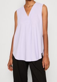 Laventeli, ilmanhihoja oleva paita, jossa on V-kotelo, rento malli ja hienovaraisia laskoksia, valmistettu sileästä kankaasta, yhdistetty mustiin housuihin.