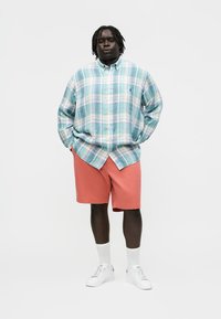 Mann steht mit Händen in den Taschen, trägt ein kariertes Hemd in Blaugrün und Weiß, korallfarbene Shorts, weiße Socken und weiße Turnschuhe.