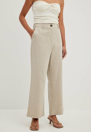 Kobieta ubrana w beżowe spodnie o szerokich nogawkach z bocznymi kieszeniami, biały top bez ramiączek oraz brązowe sandały na obcasie, stojąca na jednolitym tle.