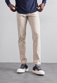 Beige maßgeschneiderte Hose in schlanker Passform, kombiniert mit schwarz-weißen Sneakers. Die Hose hat eine glatte Textur und besticht durch ein klares Design.