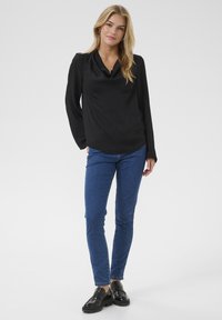 Sort langærmet bluse med draperet halsudskæring, parret med blå slim-fit jeans og sorte skinnende loafers. Enkel design, glat tekstur.