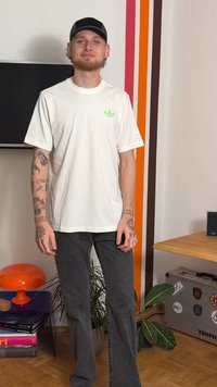 Balti medvilninė trumpomis rankovėmis palaidinė su žaliu logotipu. Derinama su tamsiai pilkomis tiesiomis džinsų kelnėmis. Foną puošia augalai ir dekoracijos.