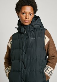 Mustat puhviliivi korkealla kauluksella ja hupulla. Varustettu vaakasaumoilla, napilla ja rintapielessä logopatchillä. Ruskea neule sen alla.