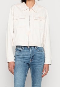 Avkortad vit denimjacka med krage, framdragkedja och två bröstfickor. Mjuk textur med en subtil blekt utseende.