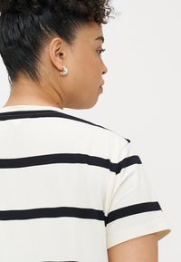 Fehér póló vízszintes fekete csíkokkal, rövid ujjal és kerek nyakkivágással. Simaszövésű pamut anyag, minimális varrási részletekkel.