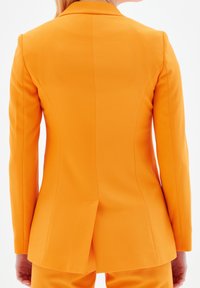 Jasnopomarańczowa marynarka o dopasowanym kroju, teksturowana tkanina, szpiczaste klapy i rozcięcie z tyłu, ukazujące krawiecką elegancję.