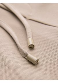 Close-up of beige textured fabric with two beige knit drawstrings ending in plastic aglets.