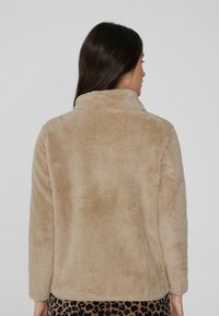 Chaqueta de piel sintética beige con un cuello alto, mangas largas y textura suave, llevada sobre una falda de estampado de leopardo negro y marrón.
