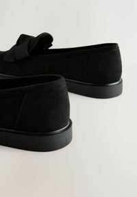 Schwarze Slip-On-Schuhe aus Wildleder mit Schleifendetail, abgerundeter Spitze und niedriger, strukturierter Gummisohle. Glatte Oberfläche mit dezenten Nahtakzenten.
