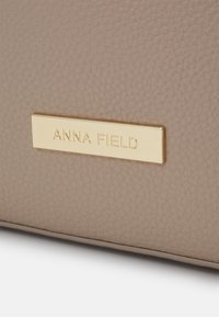 Beige struktureret læderhåndtaske med et guld rektangulært mærke prægning med teksten "ANNA FIELD." Syningerne er præcise og rene.
