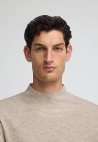 Joven con pelo corto y rizado oscuro, vistiendo un suéter beige de cuello alto, mirando al frente con una expresión neutral sobre un fondo liso.