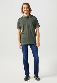 Camiseta polo verde oliva con cuello oscuro en contraste, mangas cortas y un pequeño logo. Combinada con jeans azul oscuro y zapatos negros.