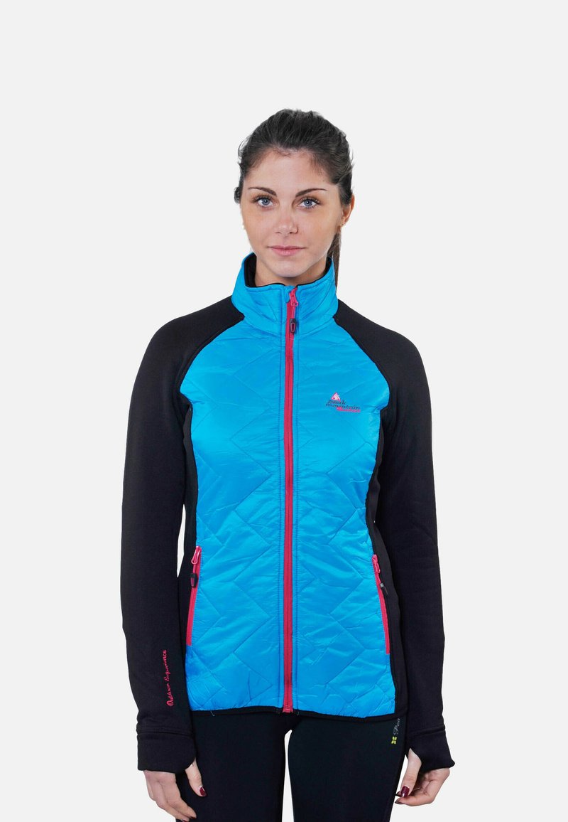 Femme aux cheveux foncés attachés, portant une veste à glissière bleue et noire avec des fermetures éclair rouges, debout devant un fond blanc uni.