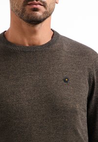 Bruine gebreide trui met een geribbelde ronde hals. Kenmerkt zich door een hexagonaal zwart logo-patch op de borst. Zachte textuur en getailleerd ontwerp.