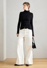 Pull col roulé noir à côtes, associé à un pantalon blanc à jambes larges, une ceinture noire et un petit sac à main noir, sur un fond neutre.