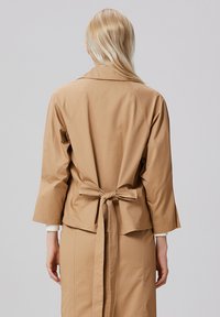 Veste légère beige avec un lien à l'arrière, des manches trois-quarts et un col ; texture lisse et design minimal sans éléments métalliques visibles.