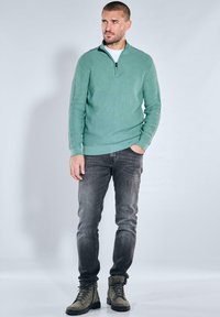 Gestrickter grüner Pullover mit Reißverschlusskragen, kombiniert mit dunklen Jeans und grünen Stiefeln, stehend vor einem hellgrauen Hintergrund.